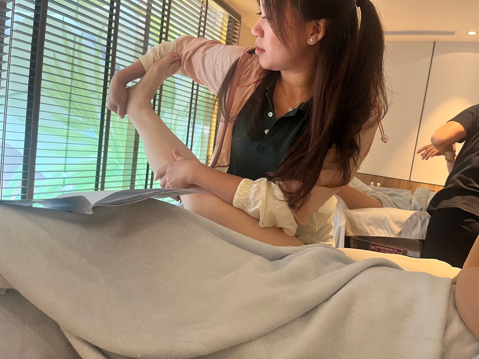 一位女士正在按摩，彎曲著手臂。按摩者躺在按摩床上。房間光線明亮，呈綠褐色調，窗簾拉上了。