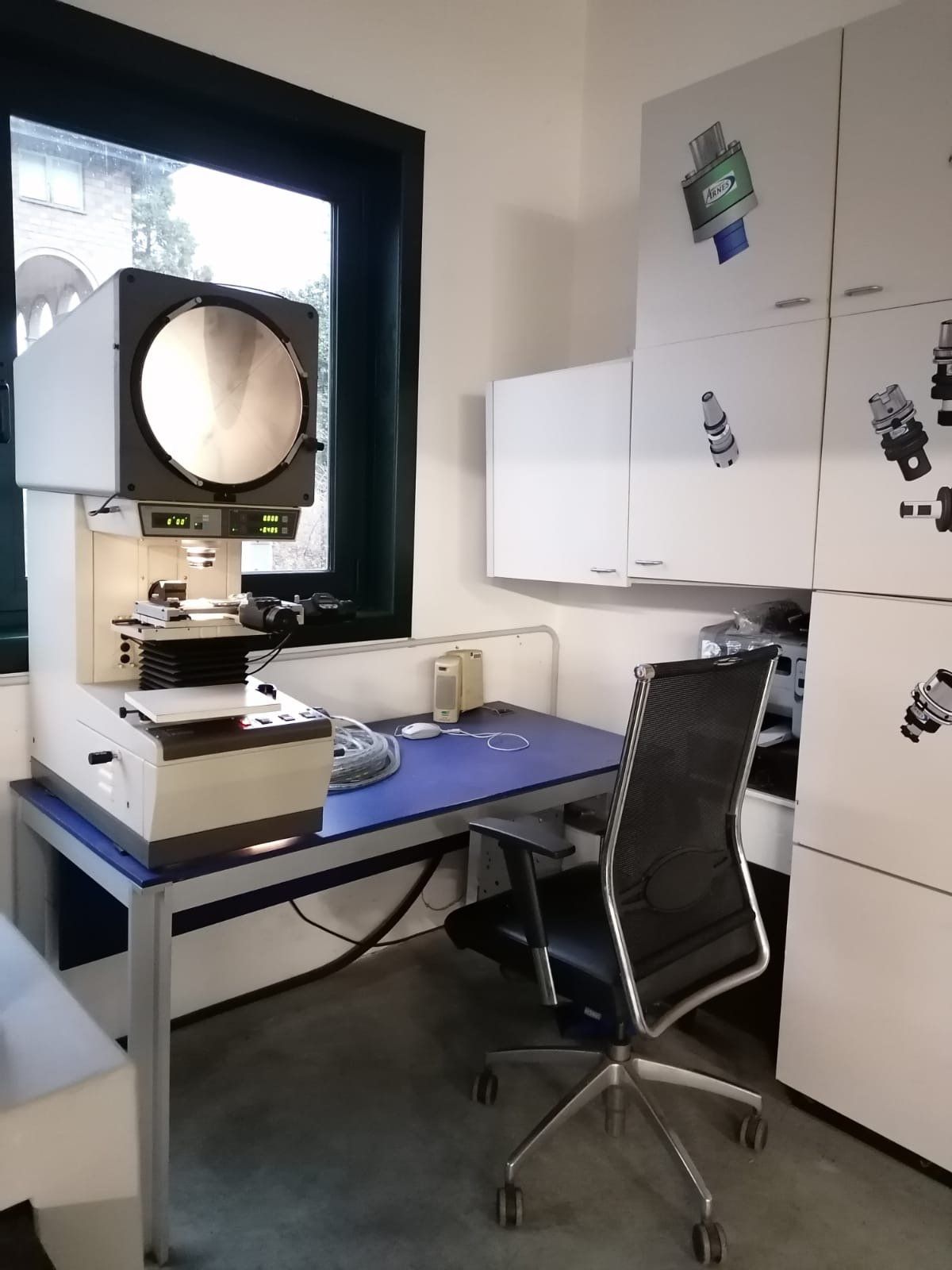 Kleines Büro mit Schreibtisch, schwarzem Stuhl, Schränken und einer großen Kamera oder einem Scanner am Fenster.