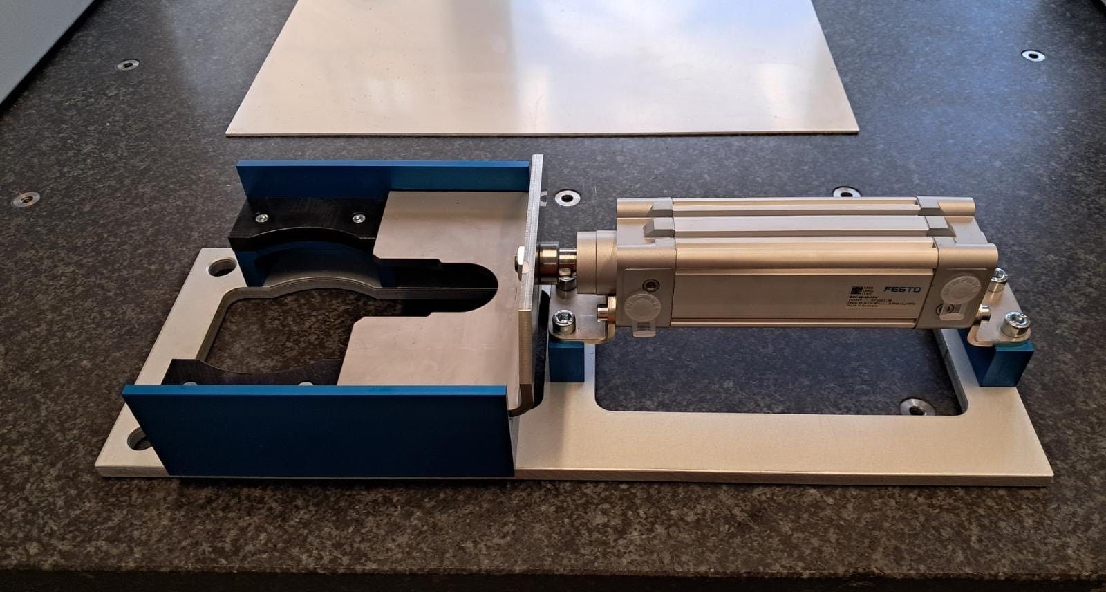 Mechanische Metallbaugruppe auf einem Tisch, mit blauen und silbernen Komponenten, die mit einem Pneumatikzylinder verbunden sind.