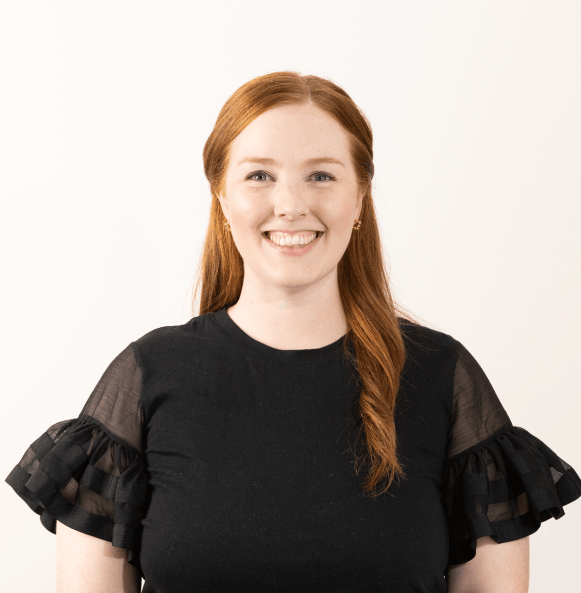 Woman with red hair smiling, wearing a black top with sheer ruffled sleeves, against a white background.