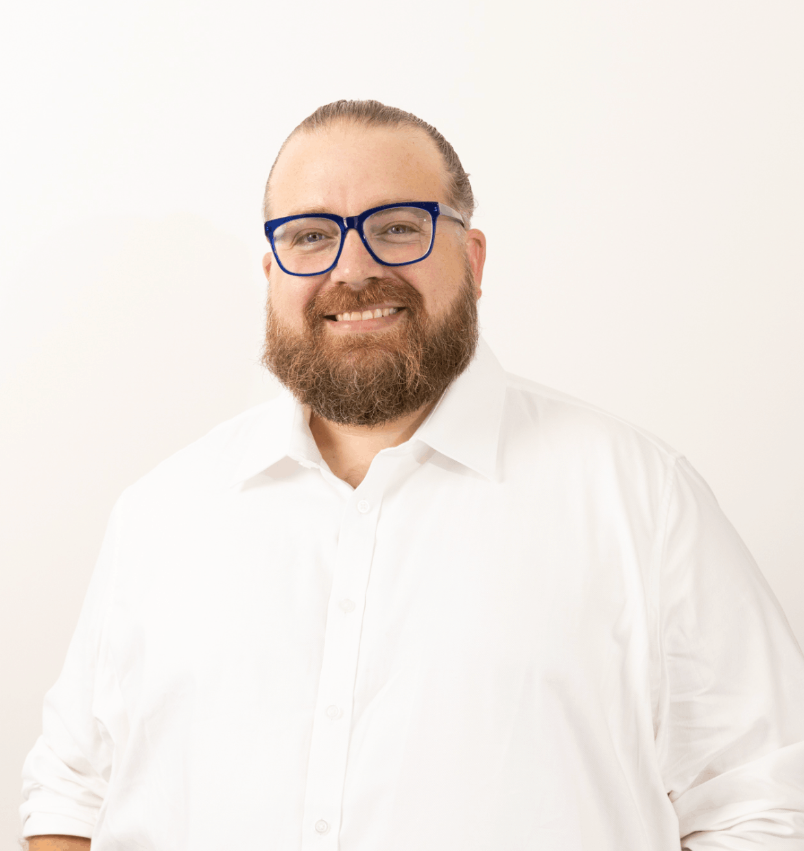 Man with a beard and glasses smiling. Wearing a white button-down shirt, set against a plain background.