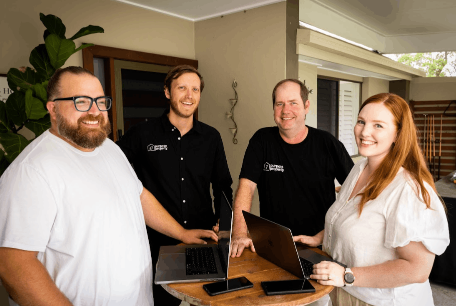 Four people smiling at a table with laptops. Outdoors, a partly shaded patio with wooden accents.