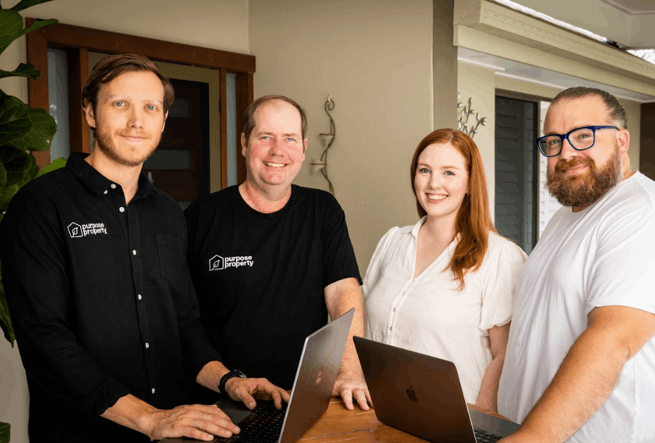 Four people standing together with laptops, smiling. Indoors, near a doorway.