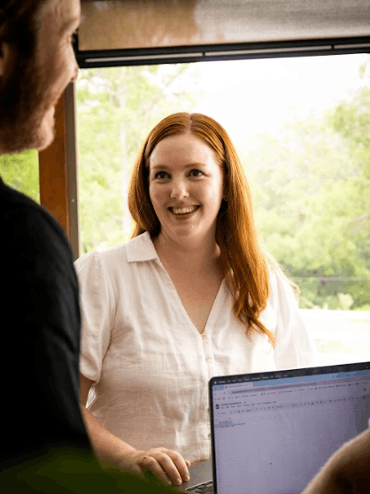 Woman with red hair smiling, talking to someone near a window, a laptop visible on the desk.