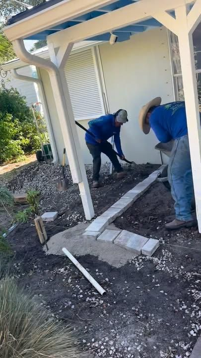 Two people in blue shirts work on a landscaping project, digging soil and laying stone pavers under a house porch.