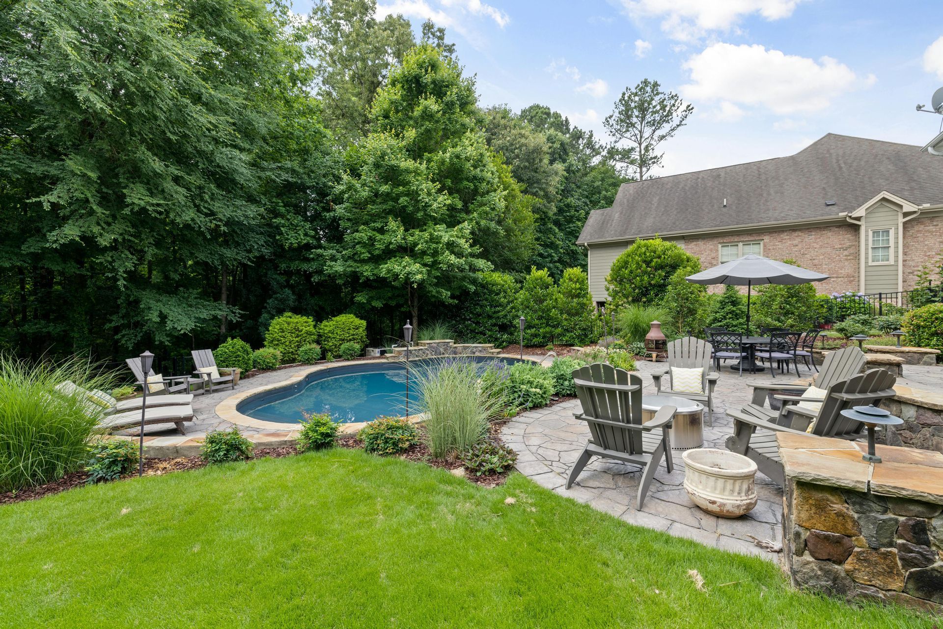 A backyard patio features a swimming pool, lounge chairs, a dining set under an umbrella, and lush trees under a blue sky.