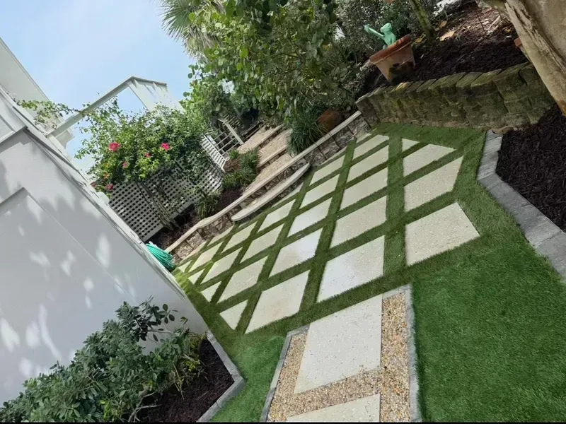 A garden patio featuring square stone pavers laid in a grid with artificial grass in the gaps, leading to stone steps.