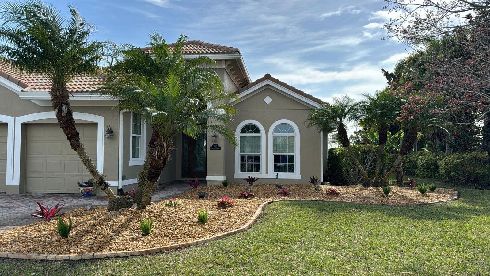 A beige, single-story suburban house with a brown tile roof, arched windows, a front garage, and a landscaped mulch bed.