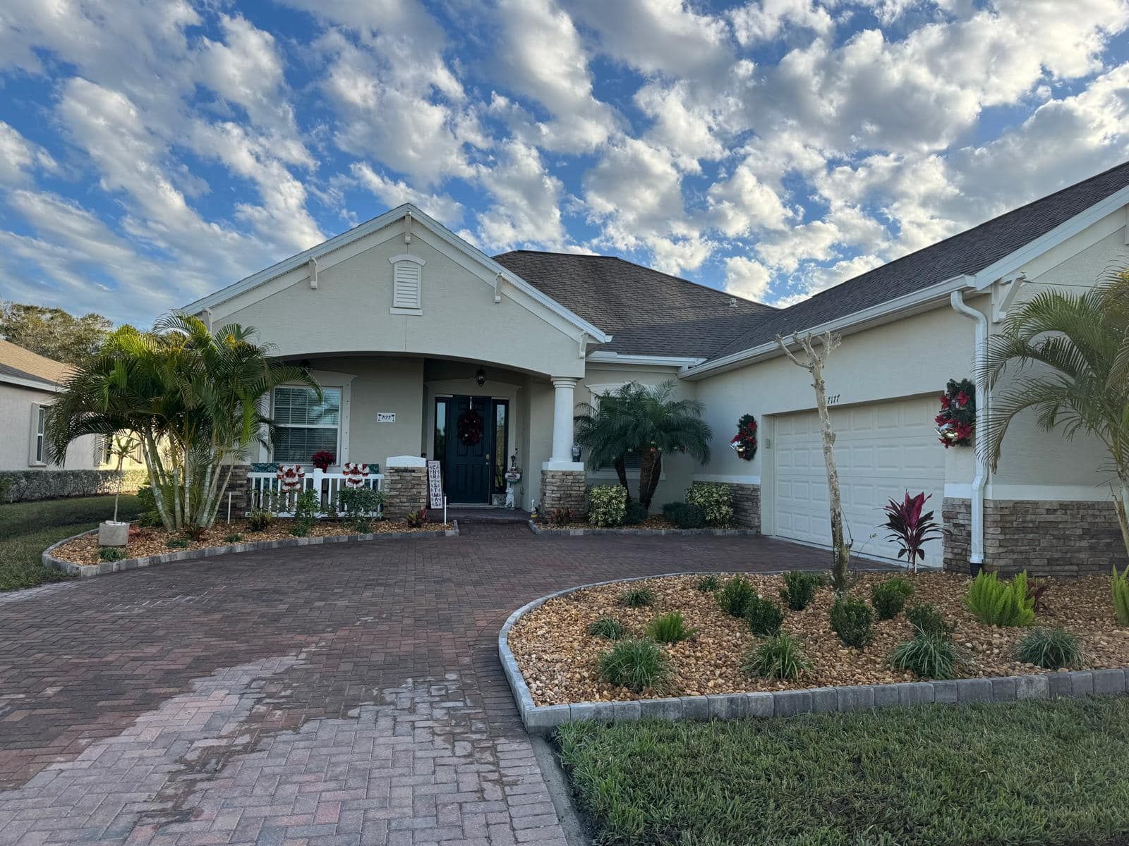 A one-story suburban home with a paved driveway, light-colored stucco exterior, and palm trees under a cloudy sky.