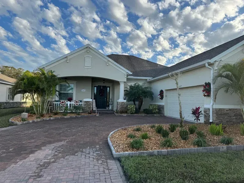 A beige single-story house with a brick driveway, palm trees, and a landscaped front garden under a cloudy blue sky.