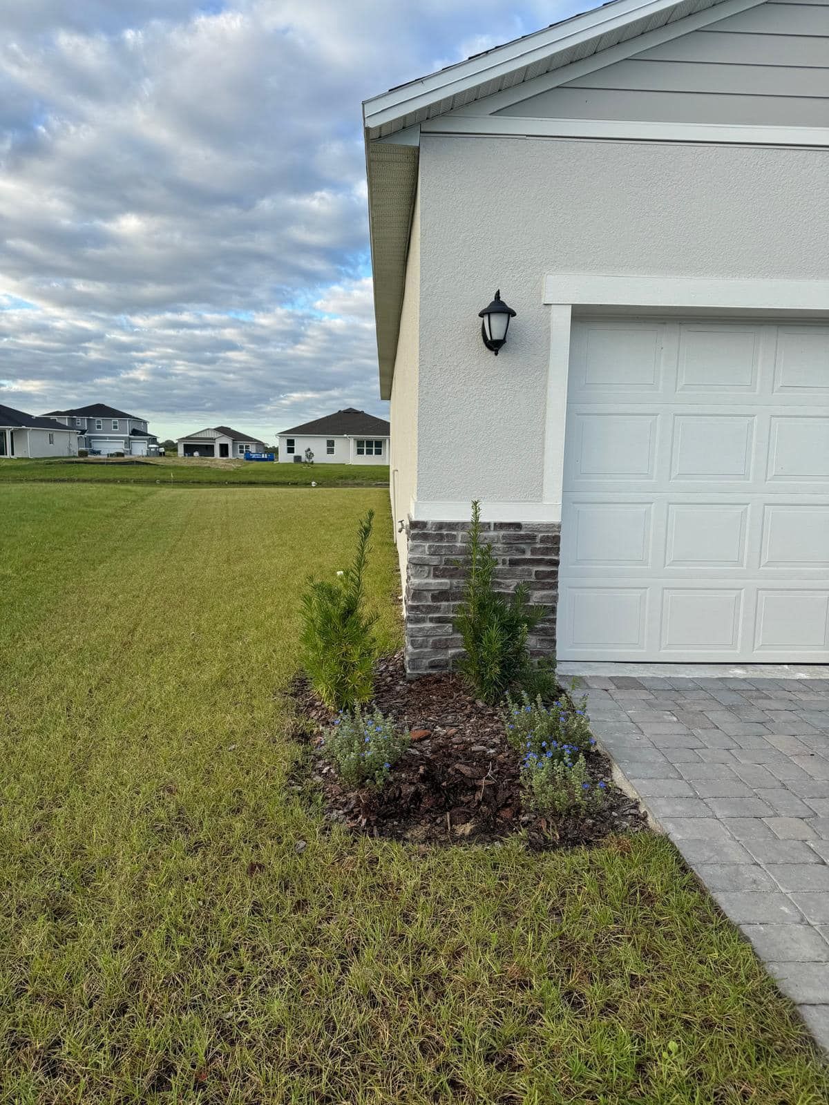 A side view of a house garage exterior featuring light stucco walls, stone accents, a wall lamp, and a small garden bed.