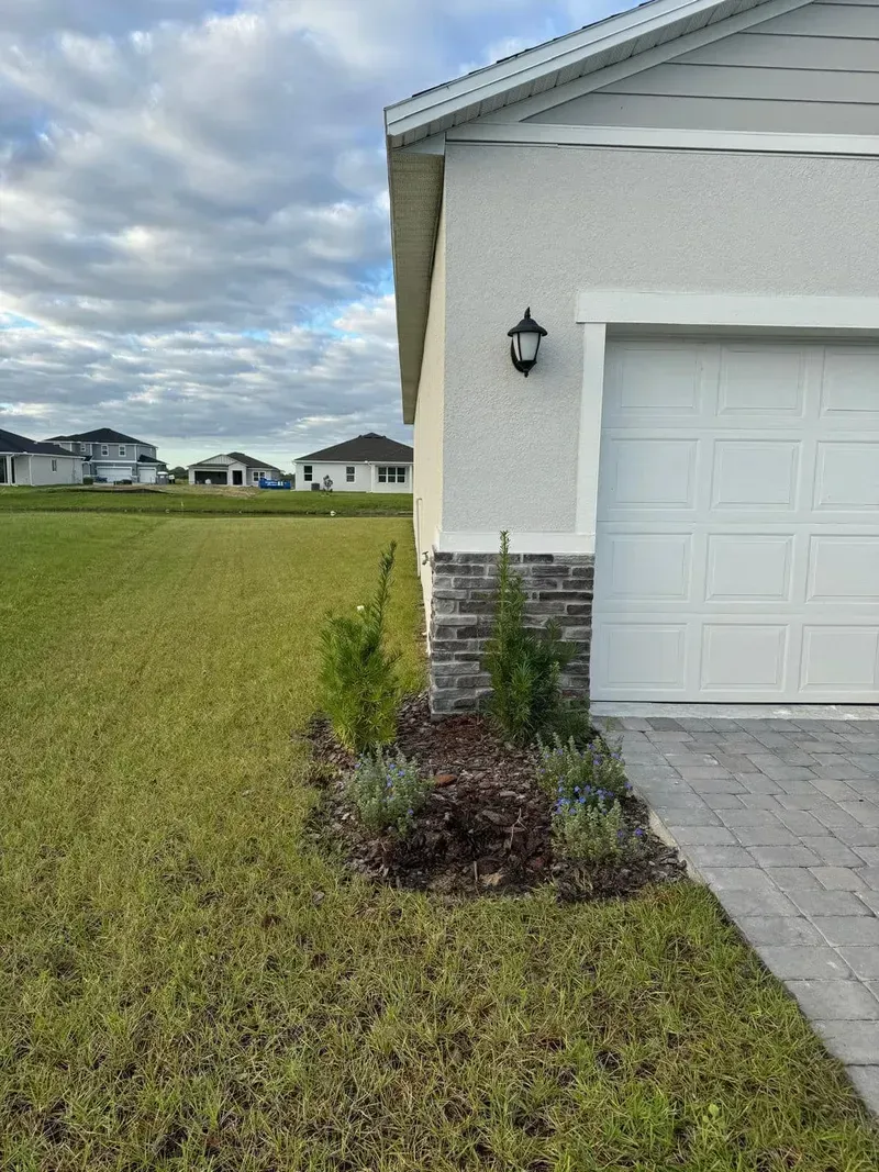 A side view of a suburban house corner with a stone base, a garage door, and a small flowerbed, next to a grassy yard.