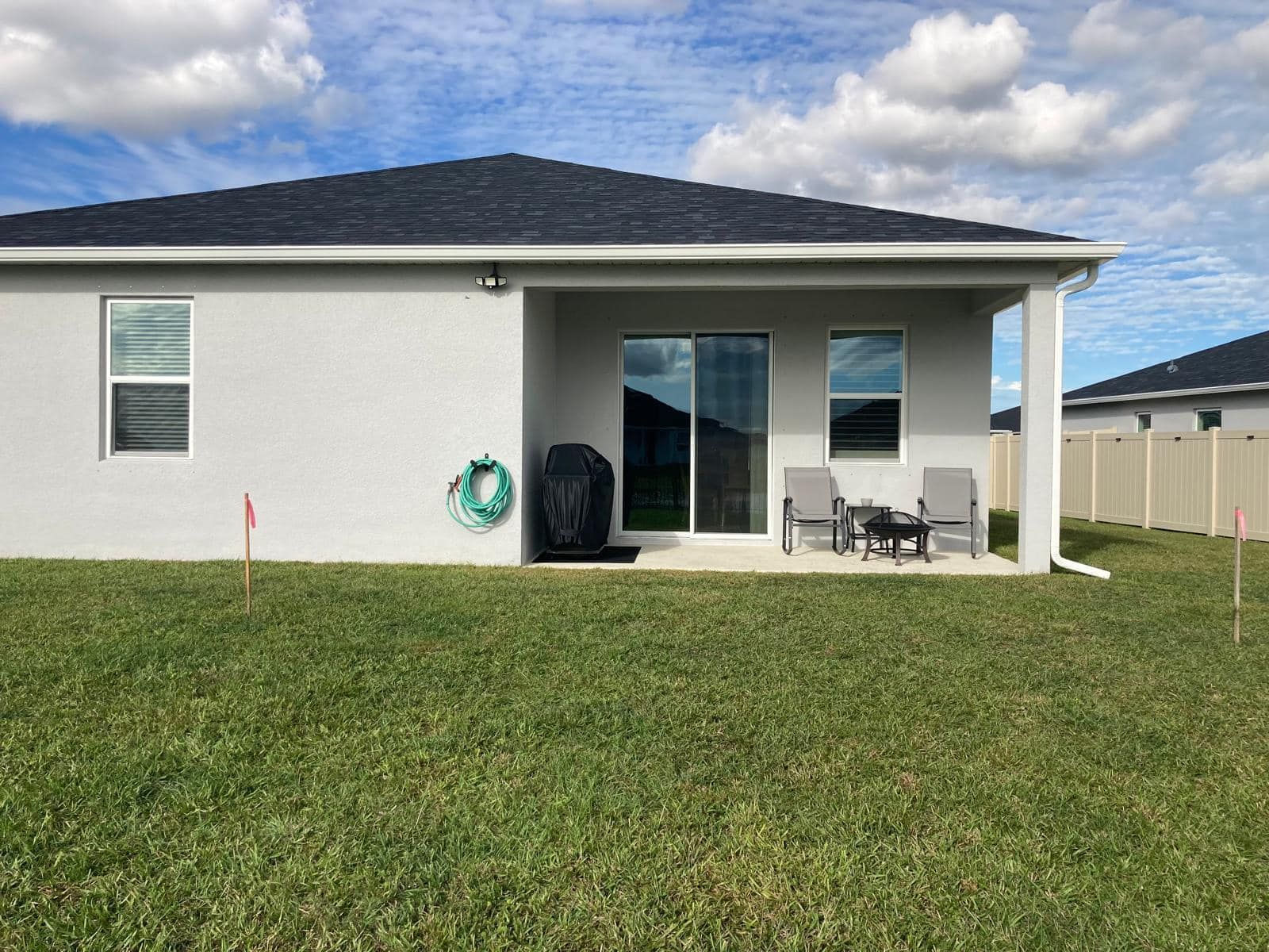 A gray house exterior with a covered patio, featuring sliding doors, two chairs, a small table, and a grill on a lawn.