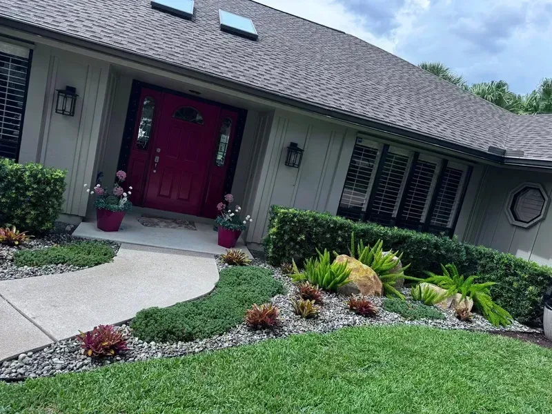 A front entrance of a home with a red door, a curved pathway, and a landscaped garden with shrubs and gravel mulch.
