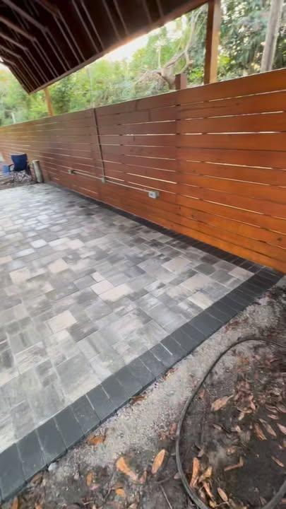A patio with grey stone pavers, a dark border, and a long horizontal wood slat fence under a covered structure.