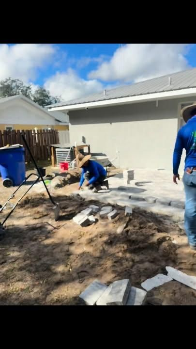 Two workers in blue shirts lay pavers on a patio area next to a house under a clear, blue sky.