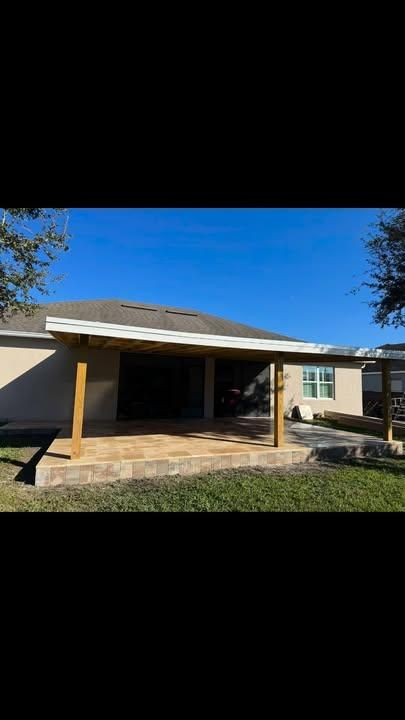 A wooden patio cover extension with a shingled roof attached to the back of a beige house over a stone-paved patio.