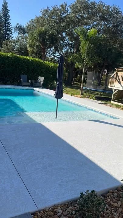 A swimming pool with a shallow tanning ledge and a closed umbrella in a sunny, fenced backyard with trees and a playset.