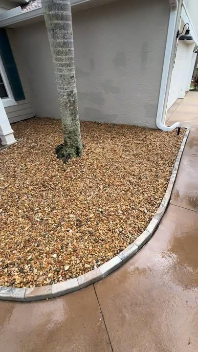 A tan pebble garden bed with a palm tree trunk, bordered by grey concrete blocks, next to a beige house and walkway.