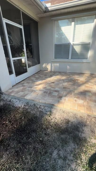 A tan stone paver patio sits outside a white house wall with a screen door and a single window.