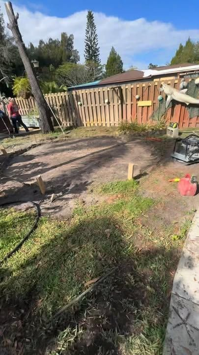 A backyard project showing a patch of cleared dirt, wooden stakes, and tools in front of a wooden fence on a sunny day.