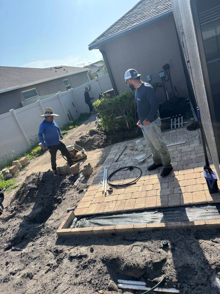 Two workers in outdoor clothing build a stone paver patio in a residential backyard next to a house wall.