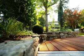 A low-angle view of a wooden deck next to a natural stone retaining wall, set within a lush, sunlit garden.