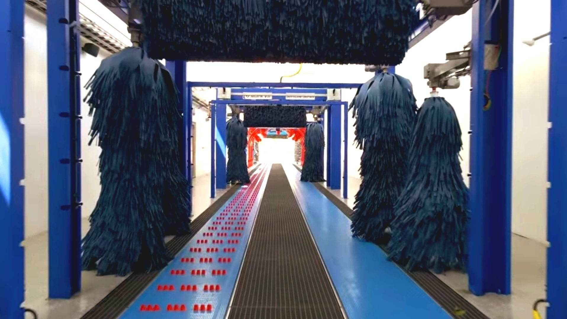 Inside of a car wash. Blue, hanging brushes line the sides and top of a tunnel. The floor has a conveyor belt.