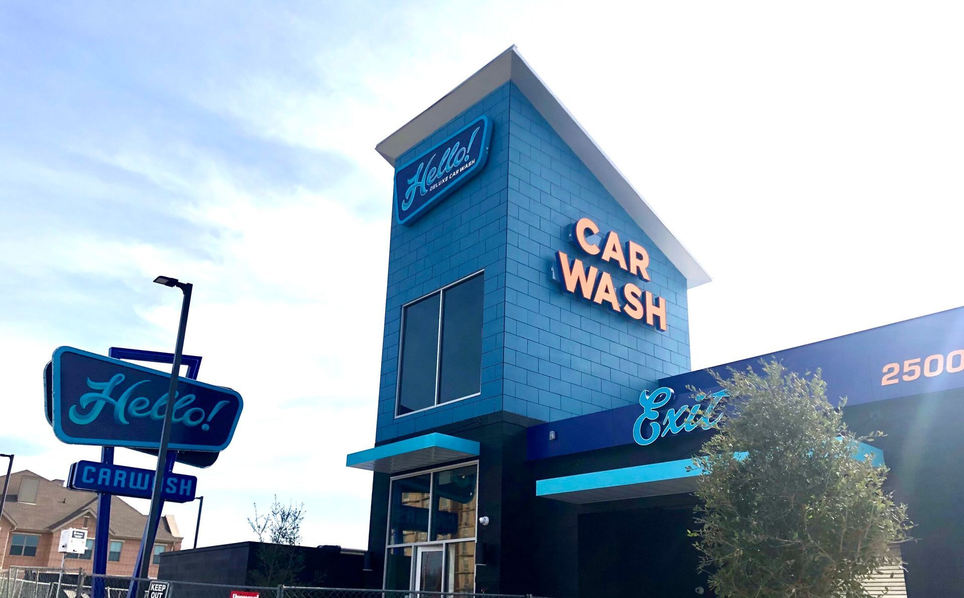 Blue and yellow Hello Deluxe Car Wash at Larmanda Street in Dallas, Tx with a white picket fence and a blue umbrella-covered table. A truck that just used the full service car wash is parked to the right.