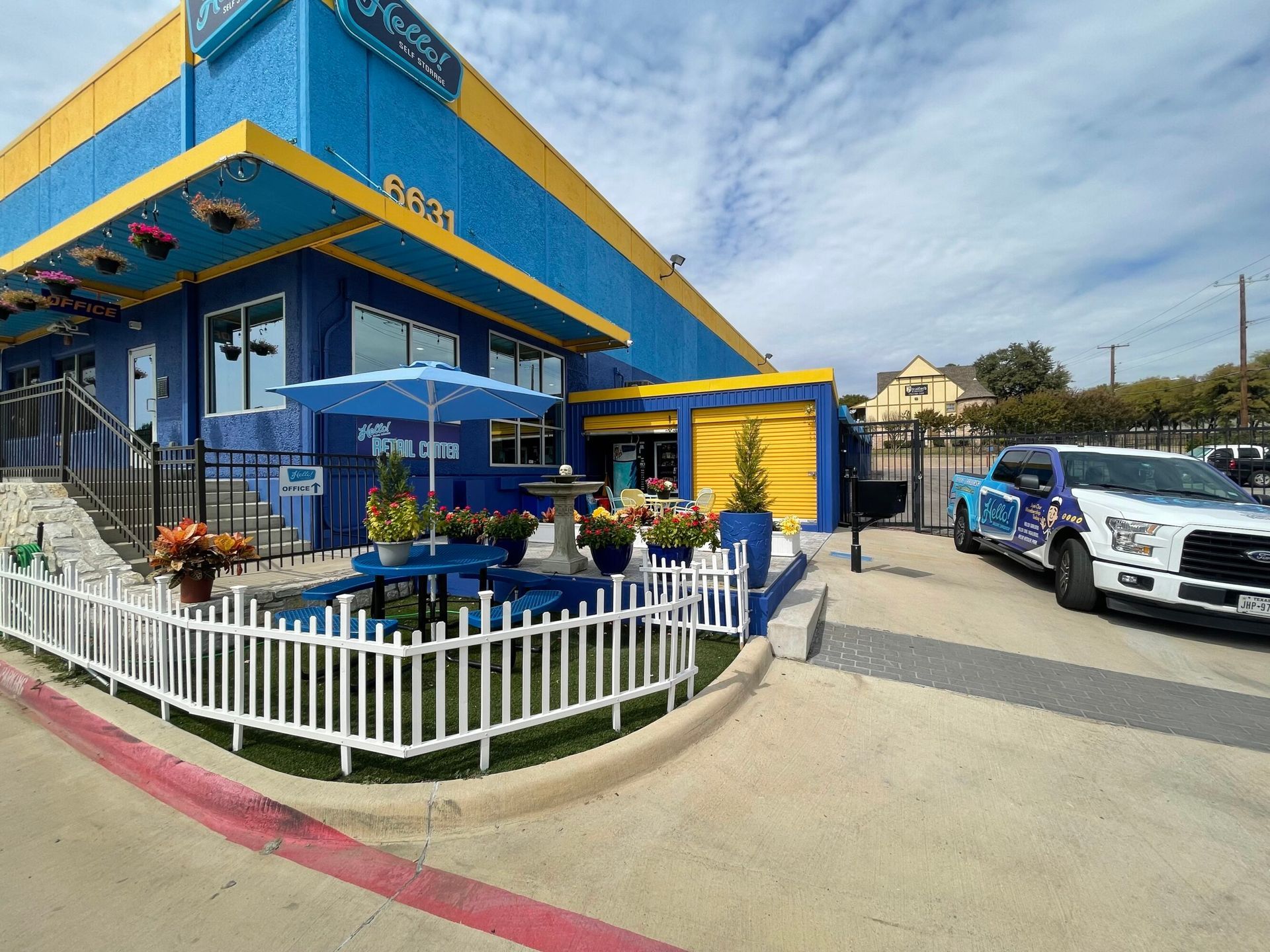 Blue and yellow Hello Deluxe Car Wash at Larmanda Street in Dallas, Tx with a white picket fence and a blue umbrella-covered table. A truck that just used the full service car wash is parked to the right.