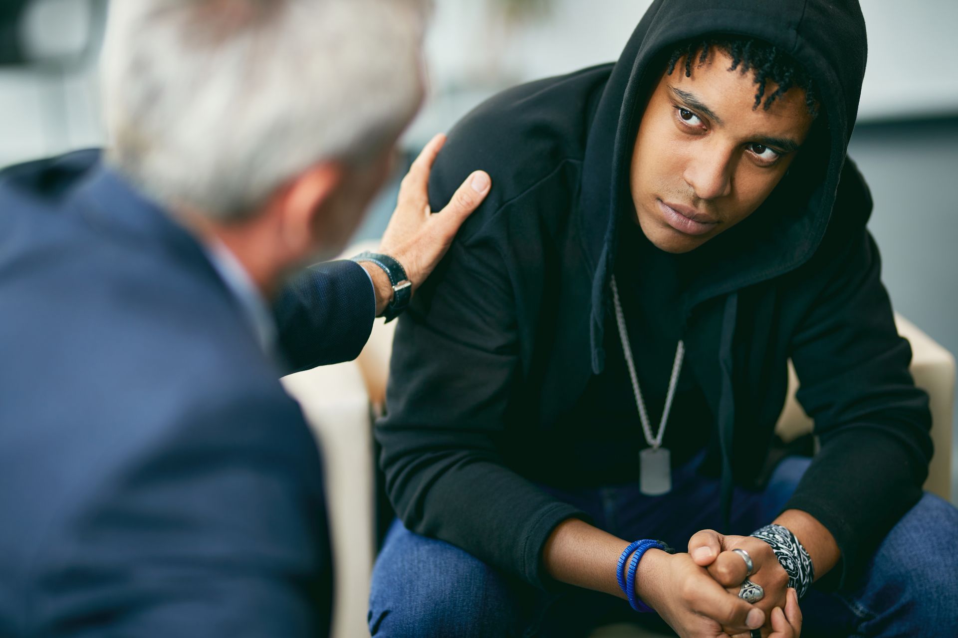 A young man in a hoodie is sitting in a chair talking to a man in a suit.