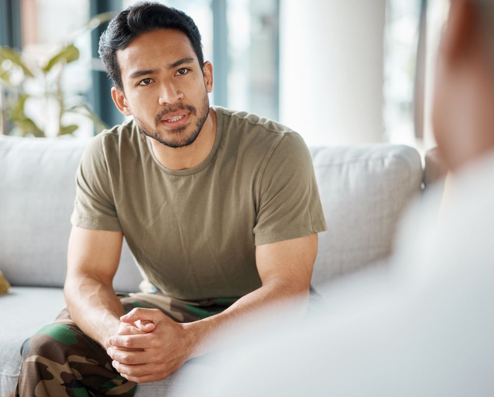 A man in a military uniform is sitting on a couch talking to another man.