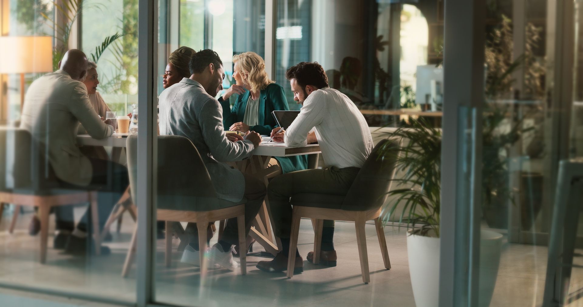 People seated around a table in a glass-walled café, chatting and drinking coffee.