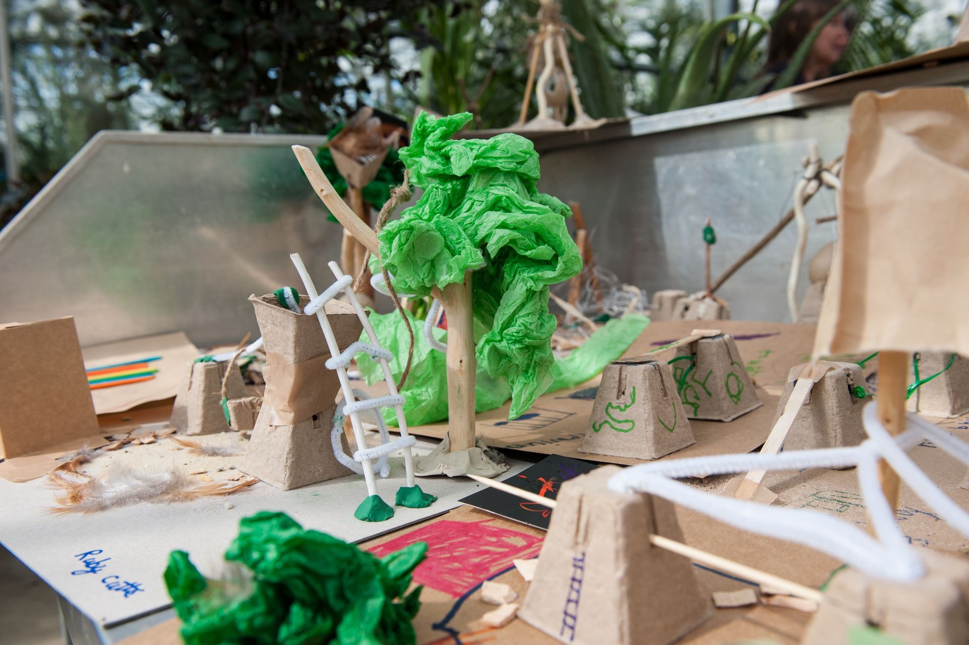 A miniature diorama with a green, textured tree and cardboard structures on a table.