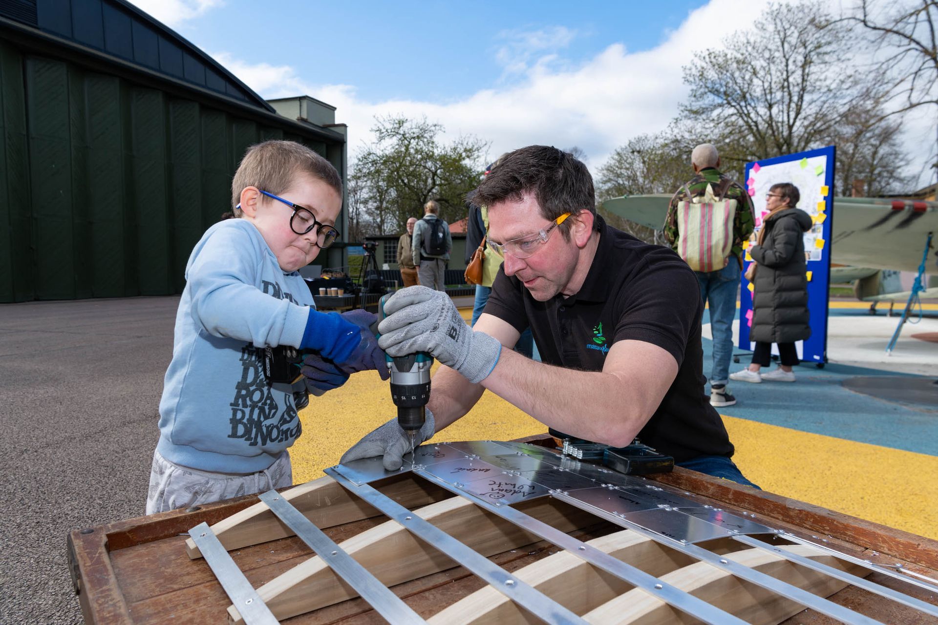 A man and a boy in glasses using a drill outdoors on a wooden frame.