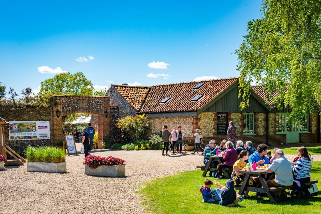 People at picnic tables outside a building with a tiled roof, under a blue sky.