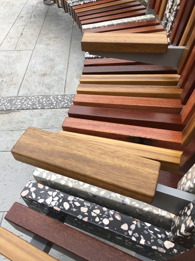 Curved bench with wooden planks of varying colors and terrazzo-like base, on a gray surface.