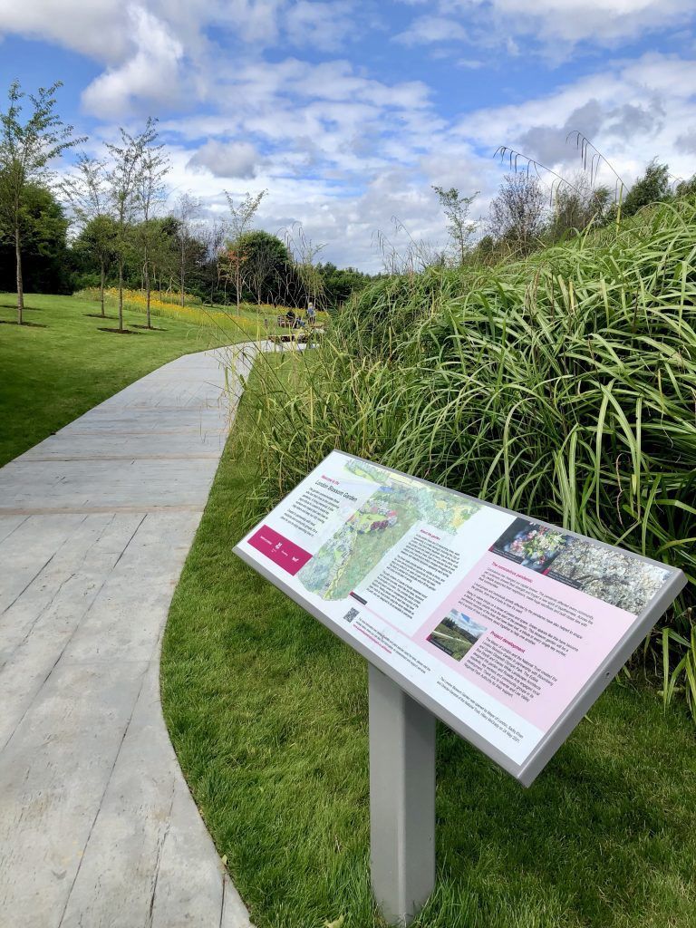 A park path leads past a sign with text, flanked by grass and green foliage, under a cloudy sky.