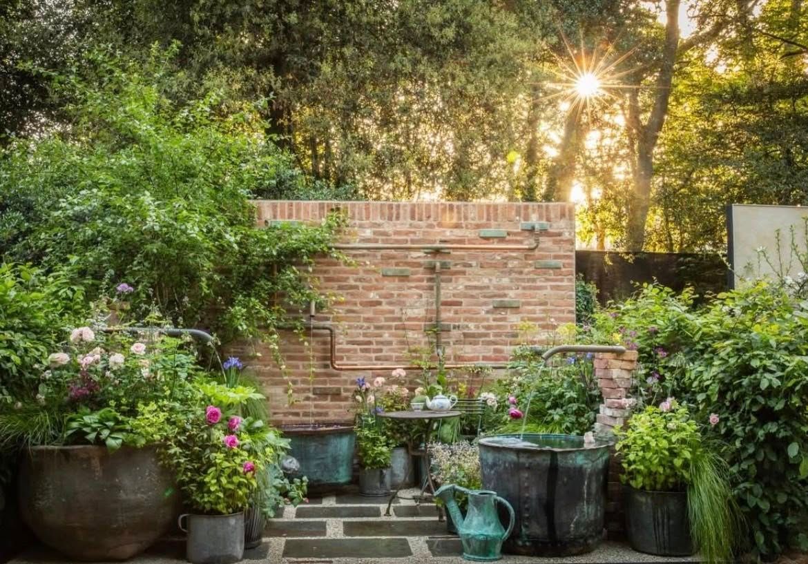 Garden pots and plants in front of a brick wall, with sunlight filtering through trees.