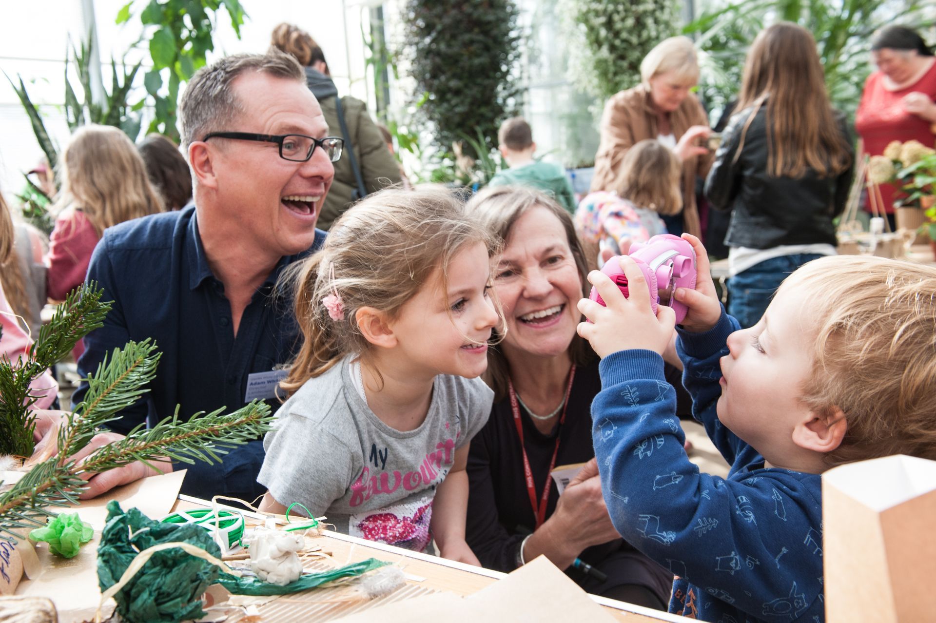 Family smiling and laughing at a craft table in a green house. A child holds a pink gift box.