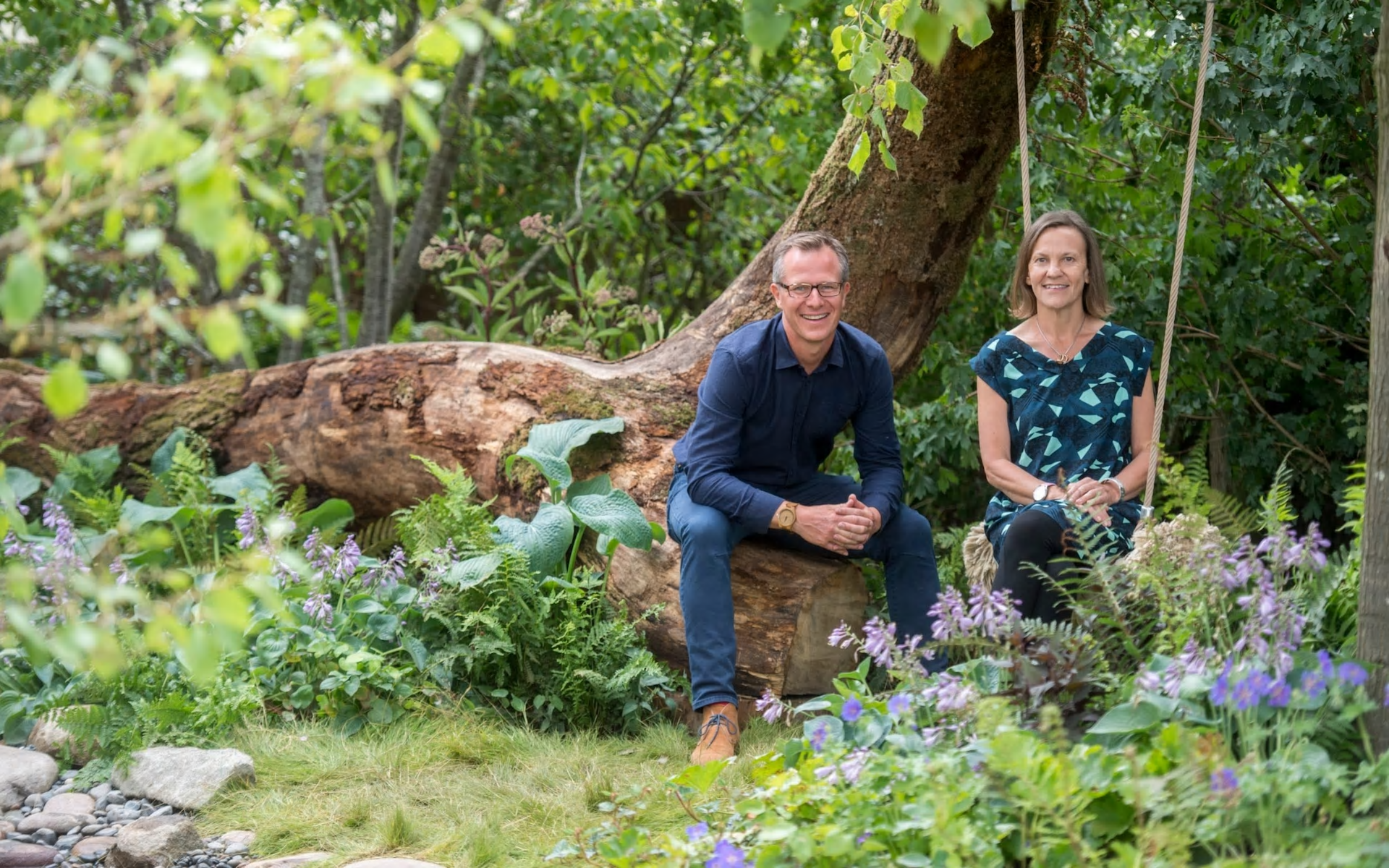 Man and woman in a garden, sitting near a fallen tree; greenery surrounds them.