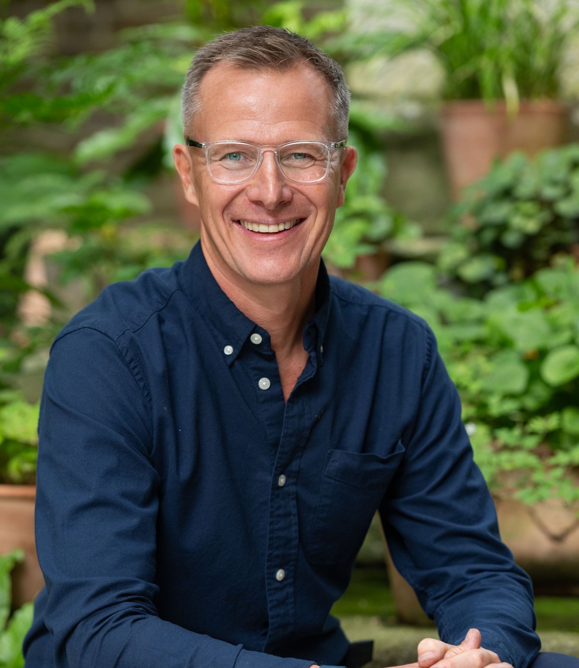 Man with glasses smiles in front of greenery. He wears a blue shirt.