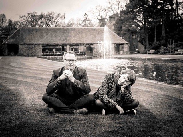 Two people sitting cross-legged on grass, in front of a pond and a building with a fountain.