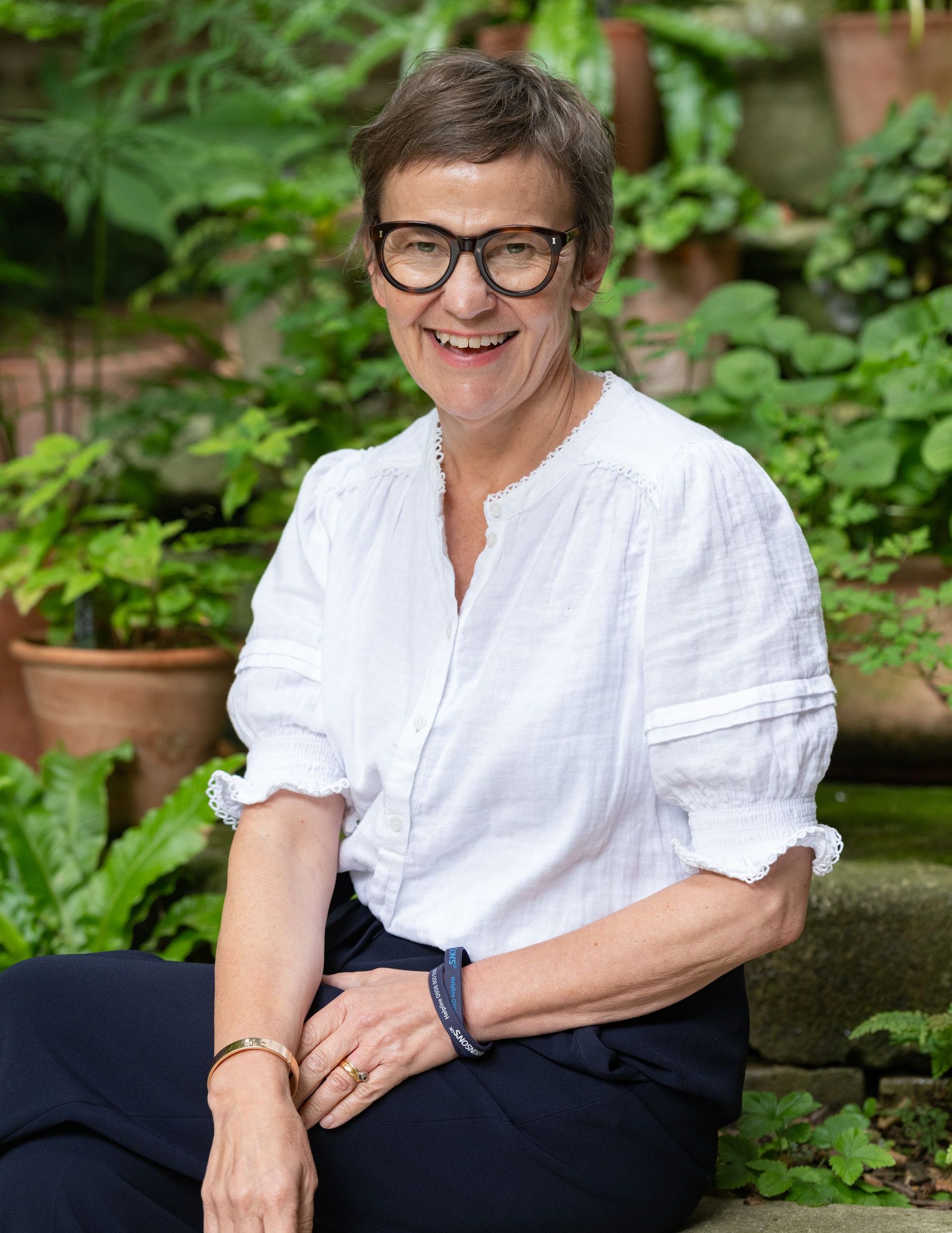 Woman with glasses smiles in a garden setting, wearing white shirt and dark pants.