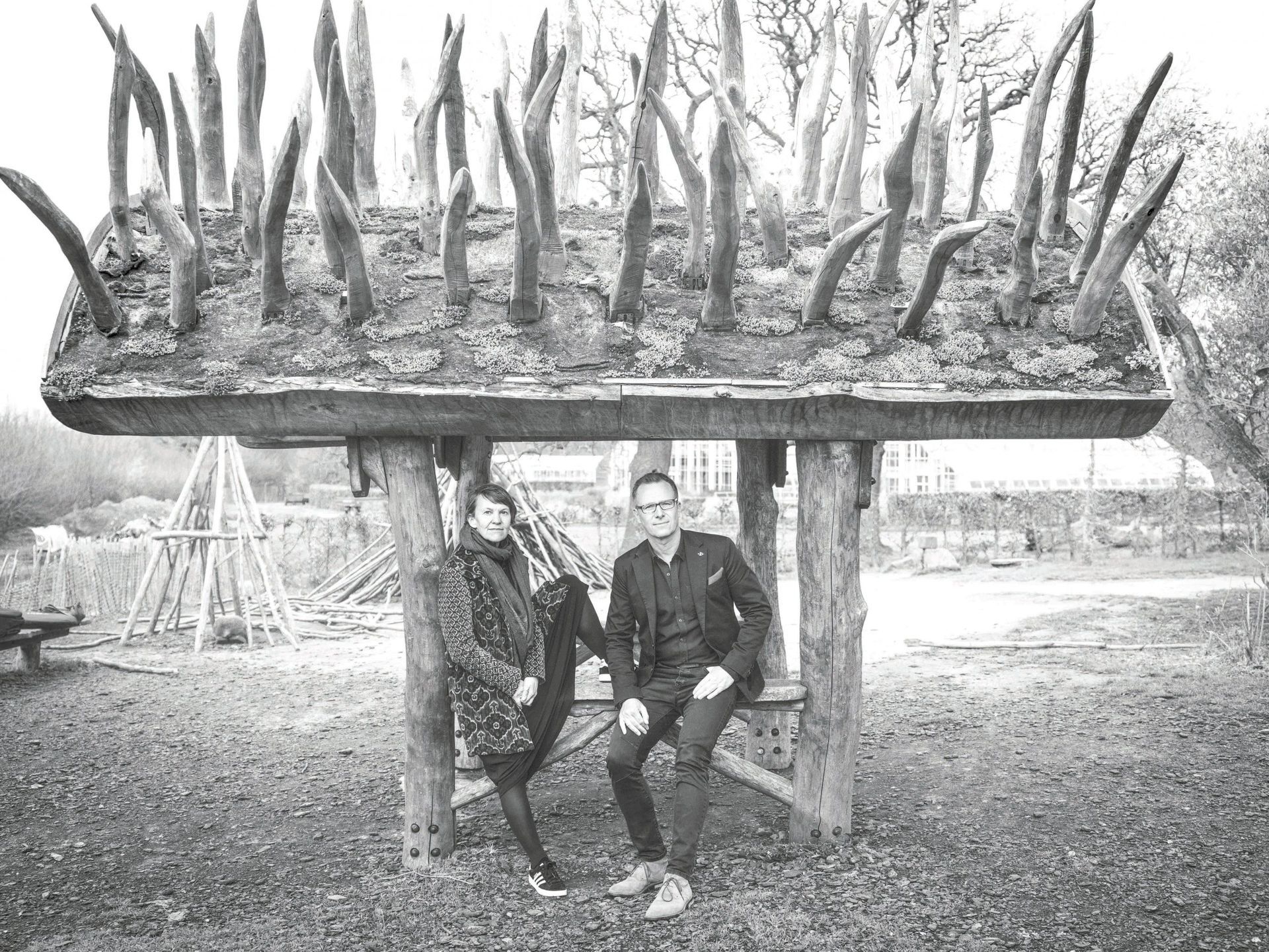 A couple sits under a wooden structure with vertical spikes. Outdoor setting, grayscale image.