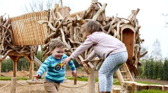 Children playing on a wooden playground; boy laughing as girl reaches out to him. Outdoors.