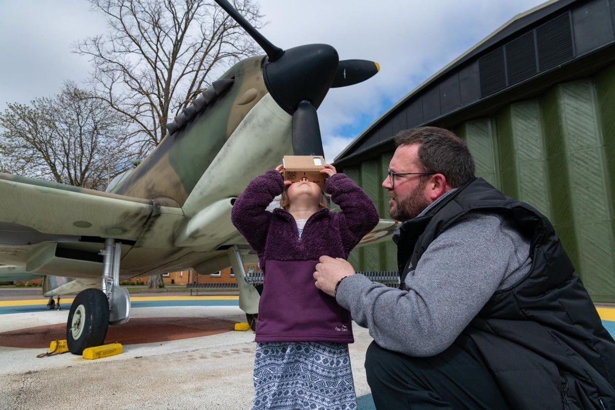 A girl with VR glasses looks up at a plane propeller, aided by a man. They're outside, near a parked warplane.