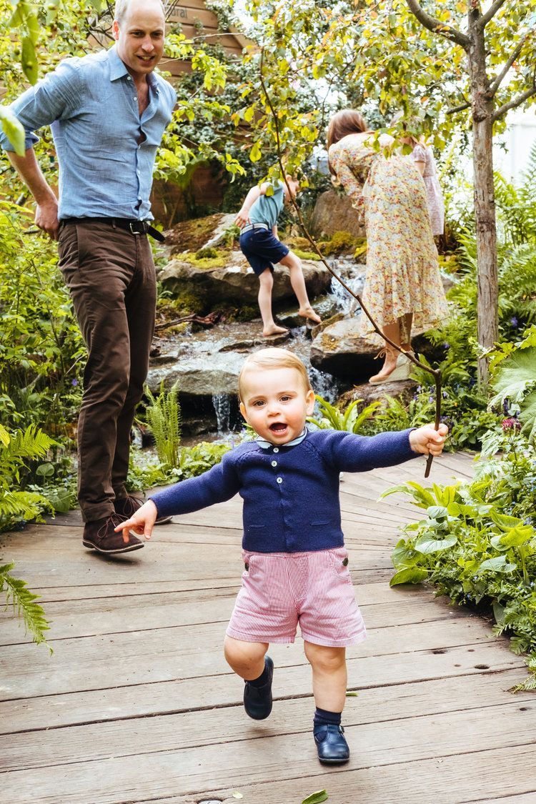 Prince George runs towards the camera, arms outstretched, in a garden, followed by his family.