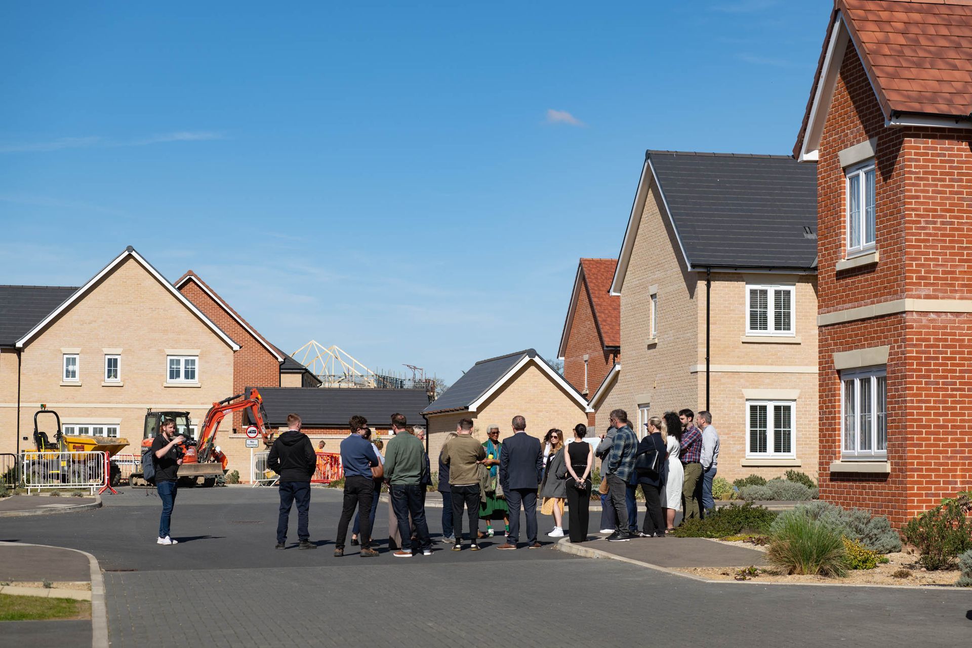 People on a street look at houses, with construction equipment visible. Blue sky.