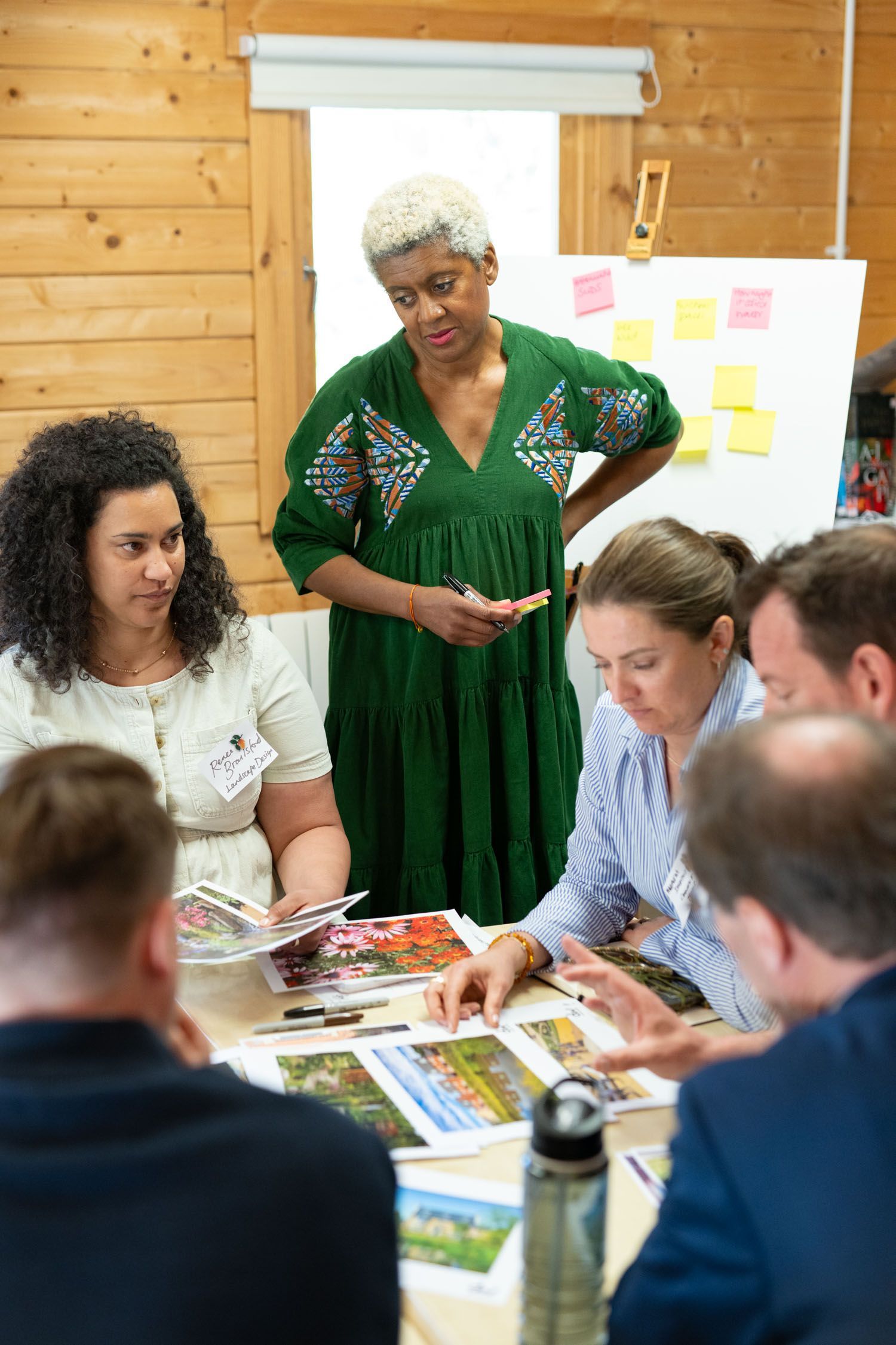 Group of people in a meeting, reviewing photographs. A woman in green dress stands, looking at the table.